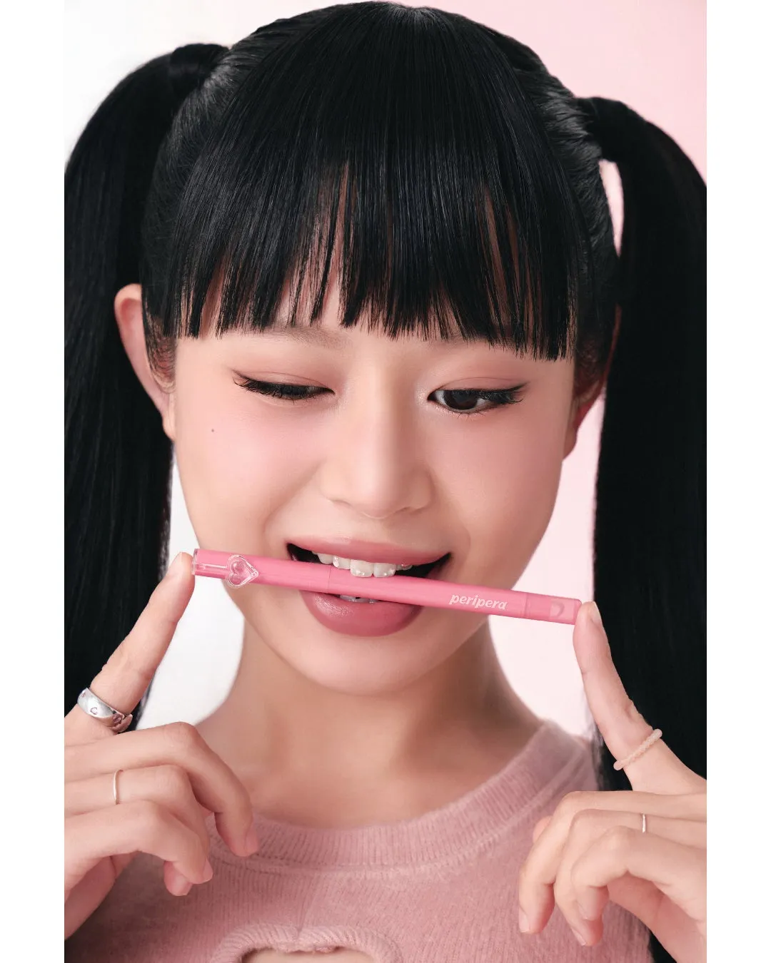 ピンク色のリップスティックを口にくわえ、指で持ち、カメラを見つめる黒髪の女性のクローズアップ。