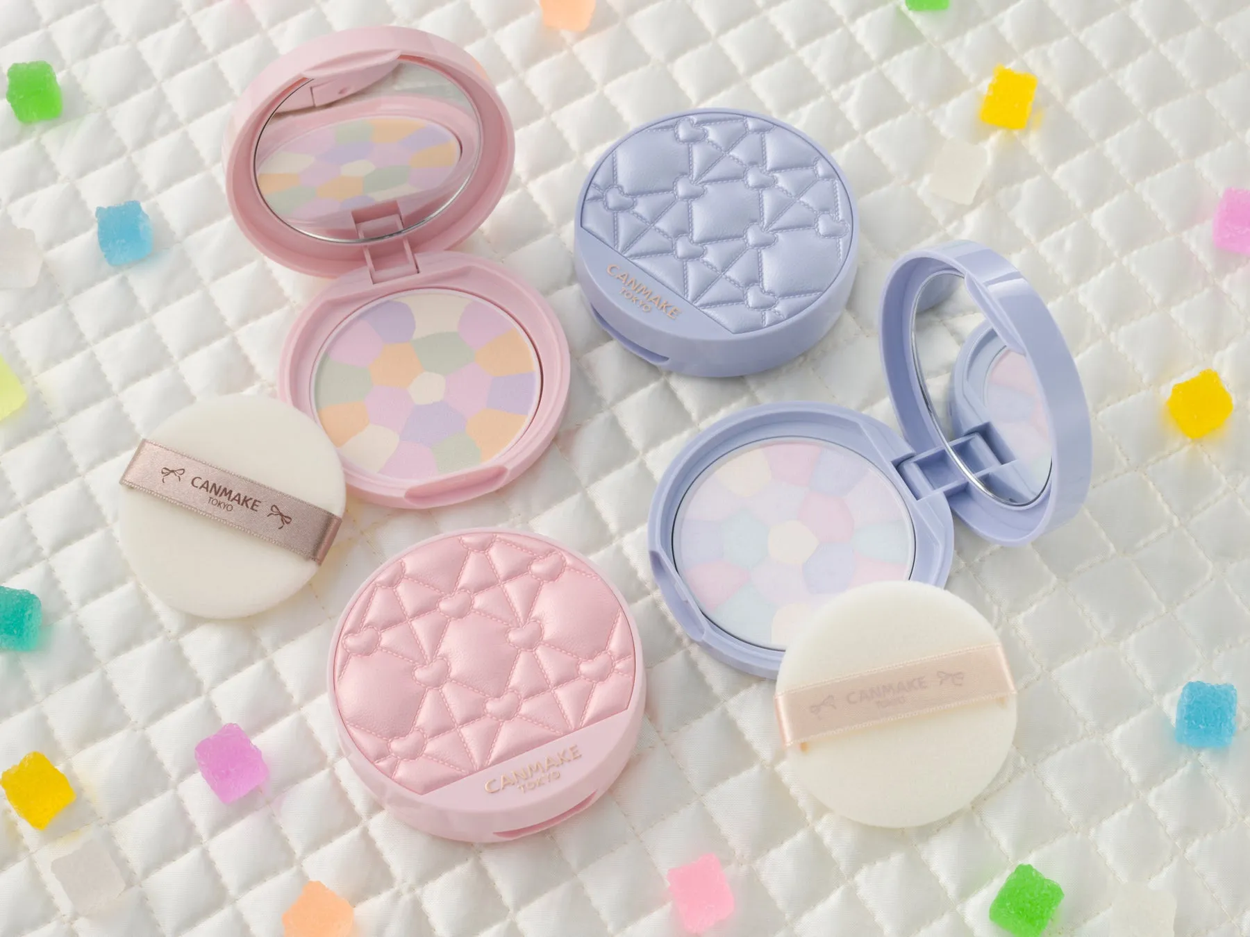 化粧品、パウダーファンデーションの製品写真です。パウダーファンデーションはピンク、水色、紫の3色で、それぞれハートやダイヤの模様が施されています。ファンデーションの横には、パフとカラフルなゼリービーンズが配置されています。