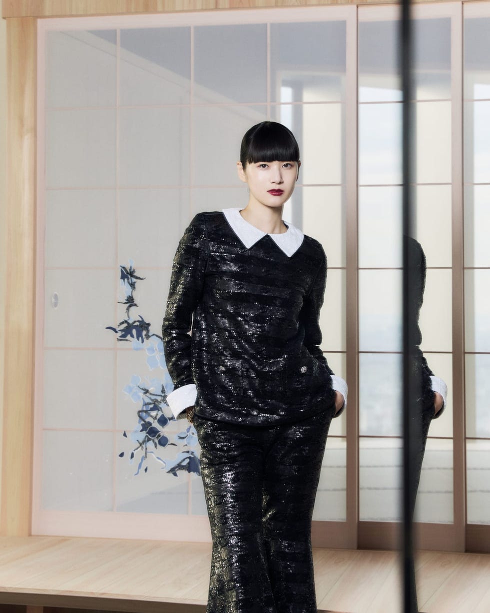 a model wearing a black textured outfit with a white collar and cuffs standing in a modern interior
