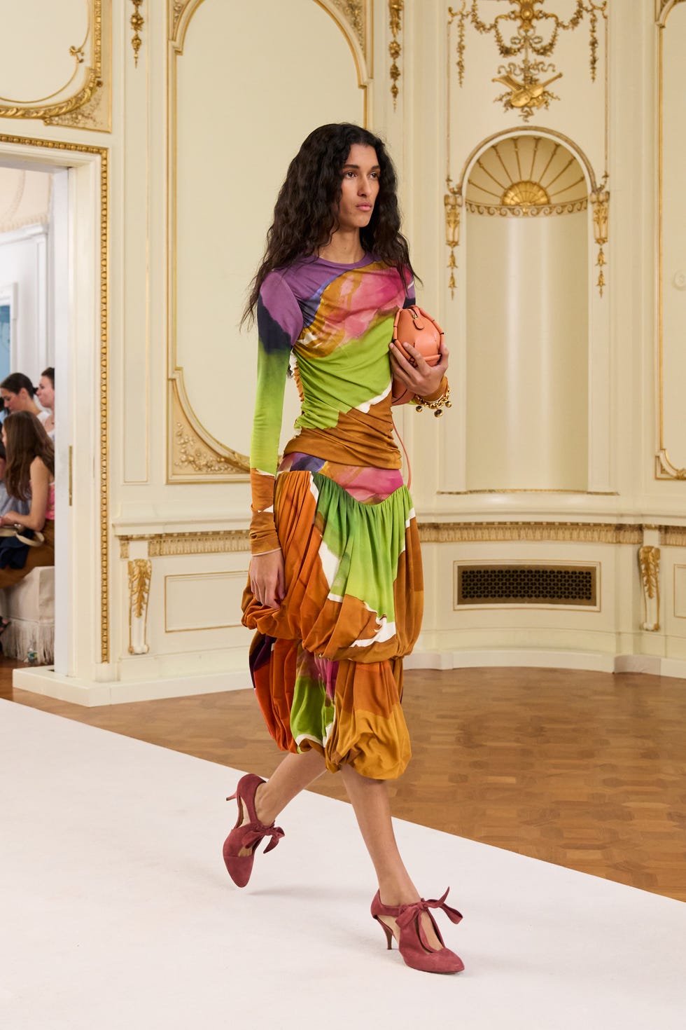 model walking on a runway wearing a colorful dress and holding a handbag