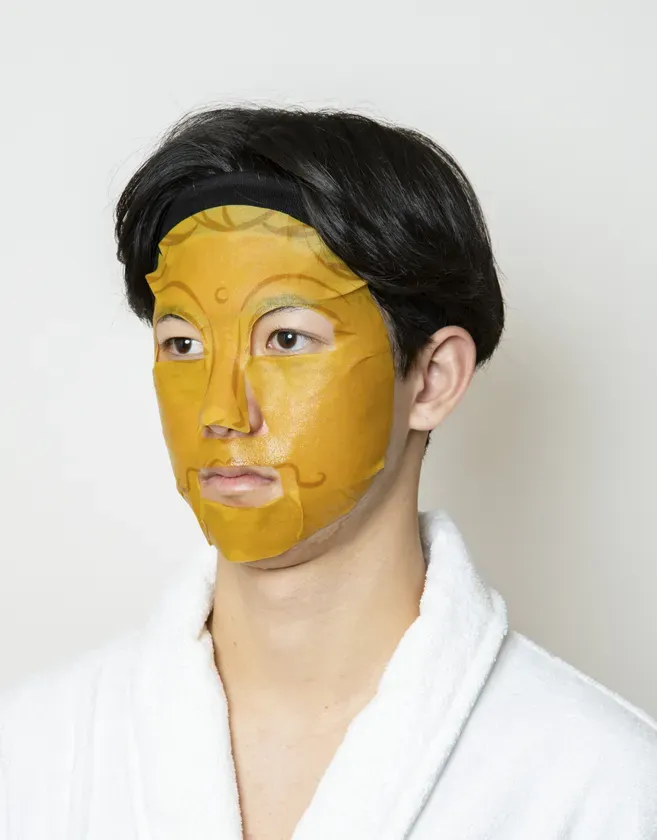 仏陀の顔が描かれたマスクを顔に装着した男性のポートレート写真です。