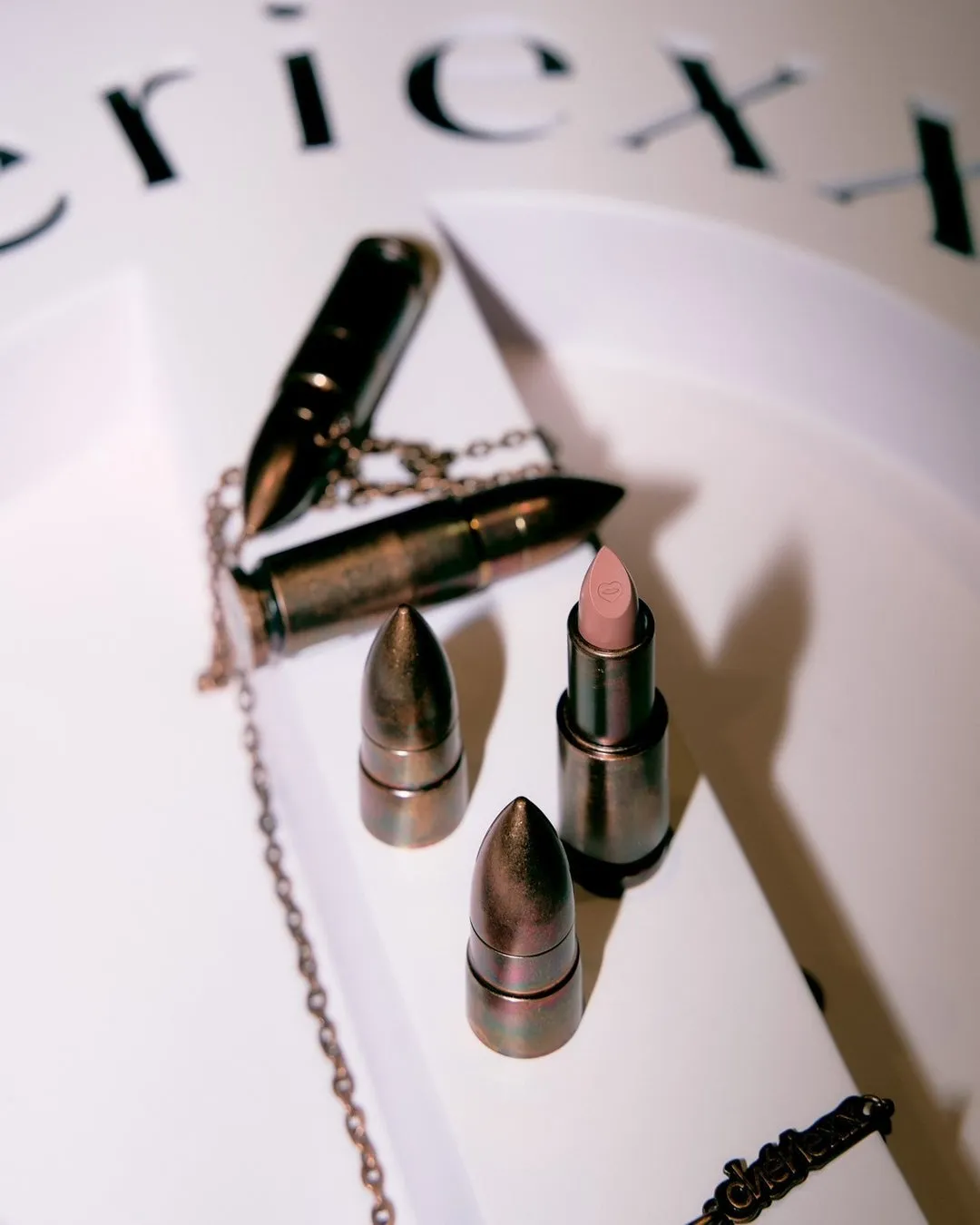 リップスティックと弾丸を模した容器が置かれた写真。リップスティックは開けられており、弾丸型の容器は装飾的なチェーンでつながれています。背景には「ericx」の文字が配置されています。