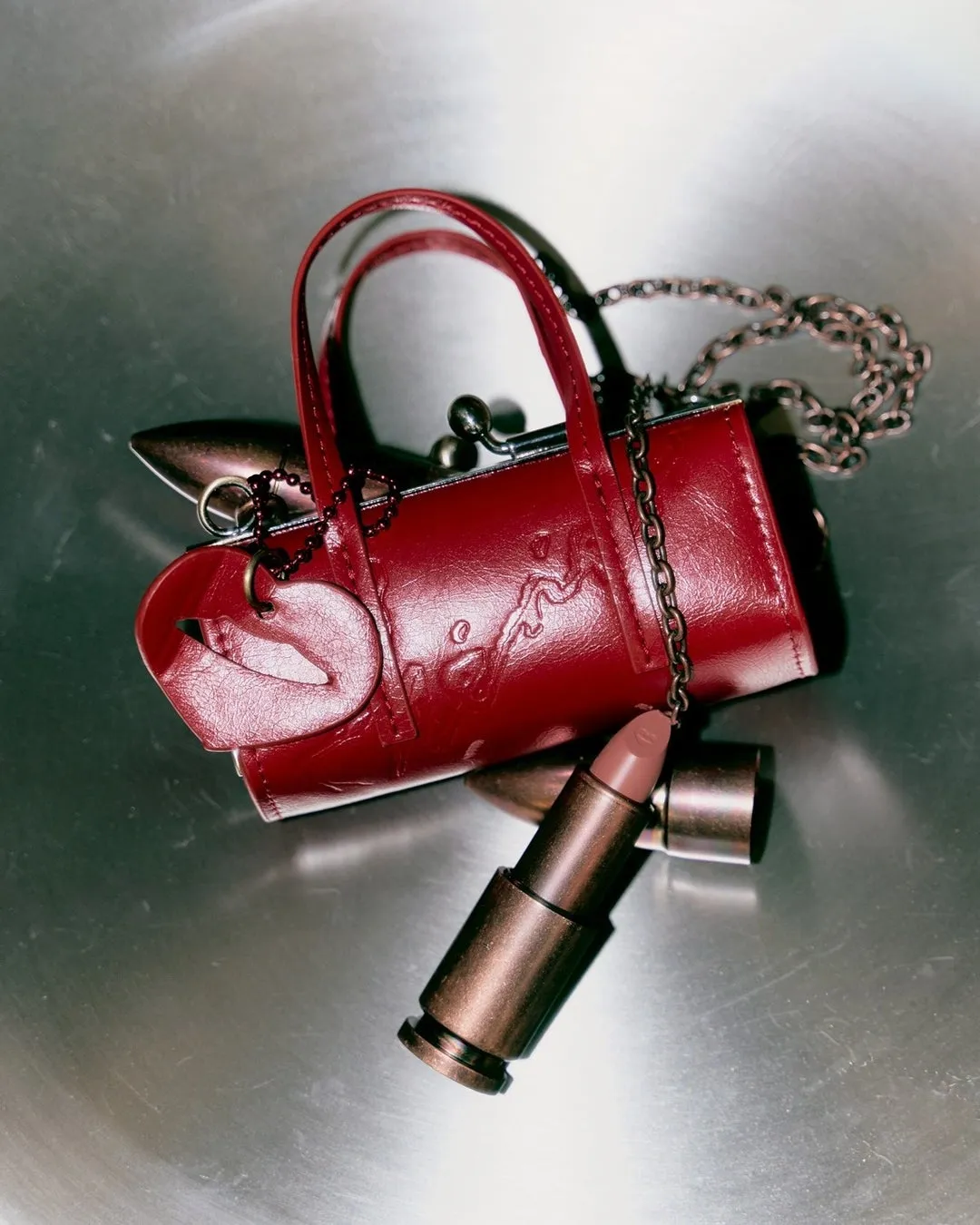 赤色のハンドバッグとリップスティックが置かれた写真。ハンドバッグには唇の形をしたチャームが付いており、チェーンが付いている。リップスティックは金属製のケースに入っている。