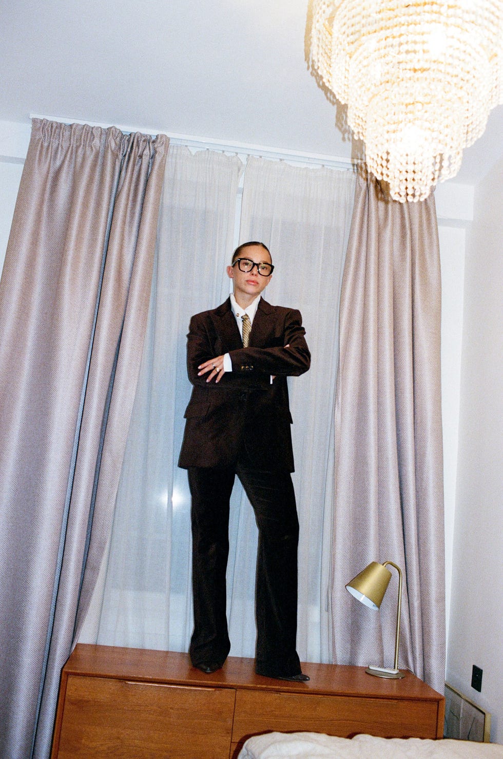 a person in formal attire standing on a piece of furniture in a room with curtains and a lamp