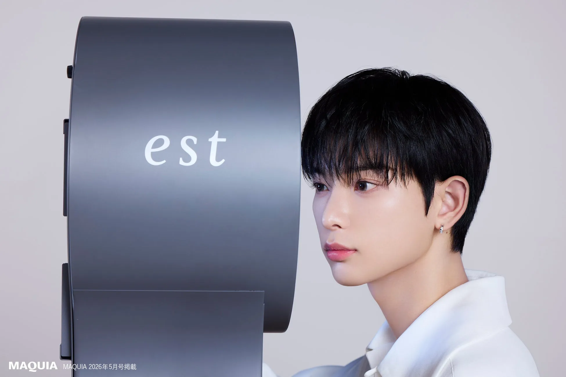 &TEAM HARUAが花王「est」スペシャルパートナーに初就任!本人コメントあり!限定ステッカー付き「砂漠スキンケア」で美肌を掴む 1 若い男性が、estと書かれた灰色のオブジェクトのそばに立って顔を横に向けているポートレート。男性は白いシャツを着ており、小さなイヤリングをしている。背景は白い。