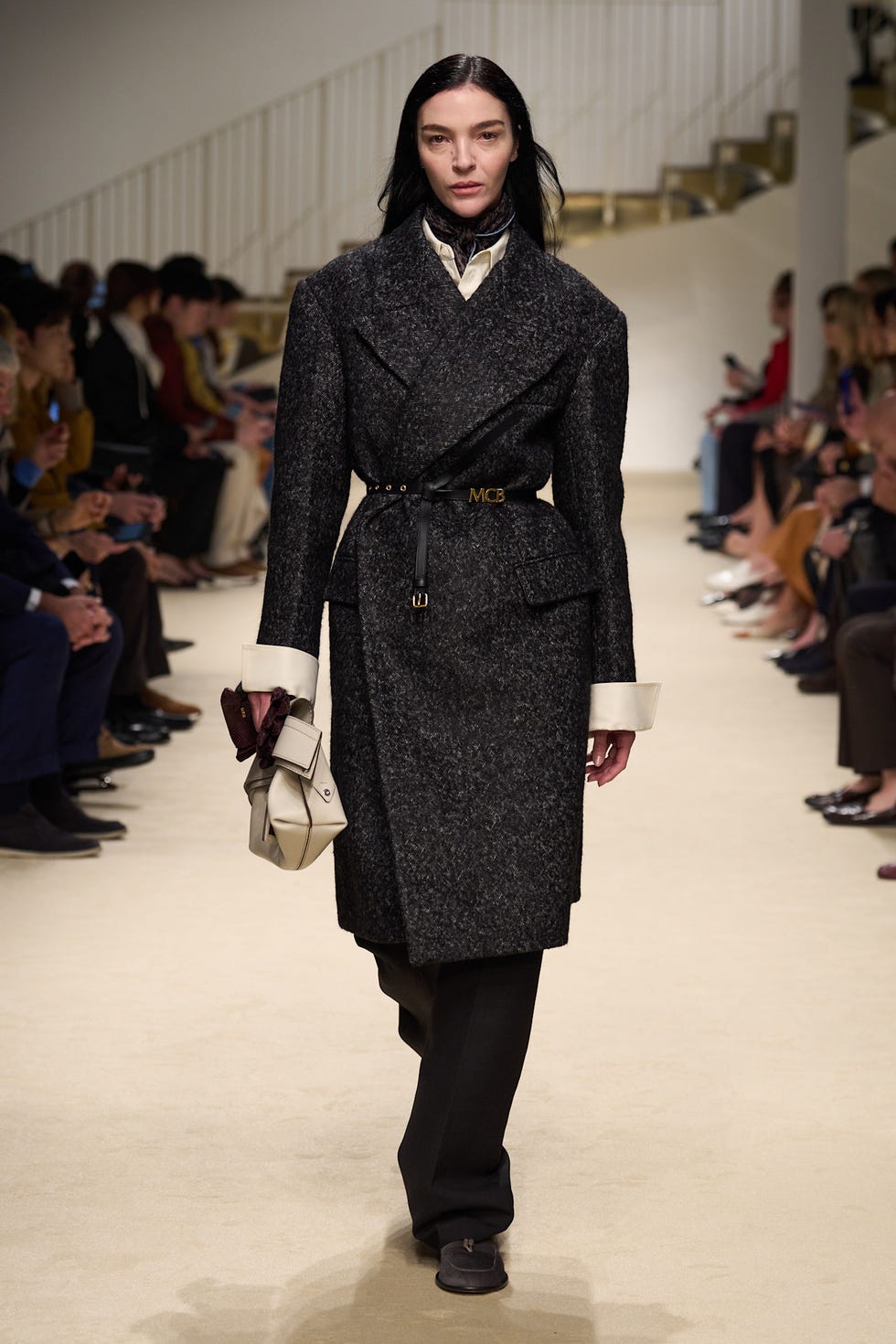 model walking on a runway in a fashion show wearing a dark coat and carrying a handbag