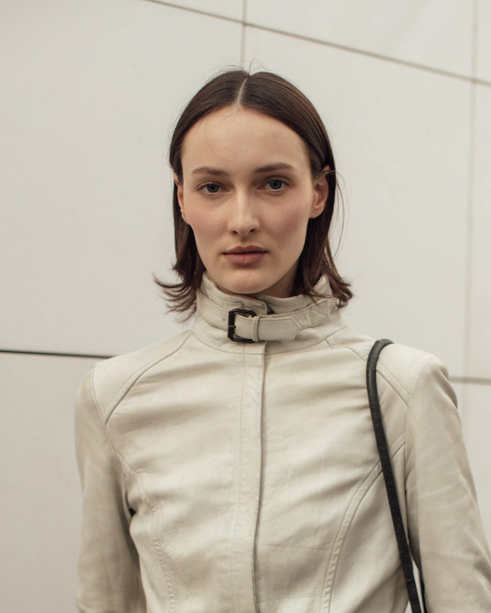 individual wearing a white leather jacket and jeans poses against a lightcolored wall